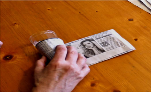 3. Enrole a tira de jornal em torno do copo, de forma que sobre um pedaço na parte inferior (ele comporá o fundo do vasinho). Coloque um pequeno pedaço de fita adesiva (a mesma será decomposta com o jornal) para fixar