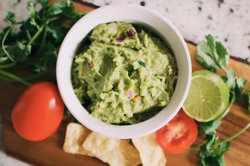 tigela de guacamole sob uma tábua de madeira contendo tomates, coentro e tortilhas. ilustra a matéria como fazer guacamole